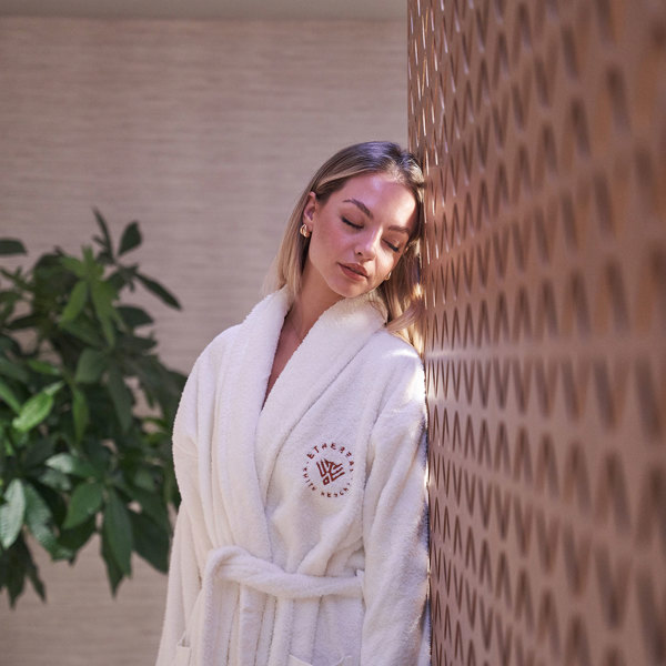 Ethereal White Resort Heraklion Spa visitor wearing a bathrobe with the hotel logo on it while laying on the wooden wall